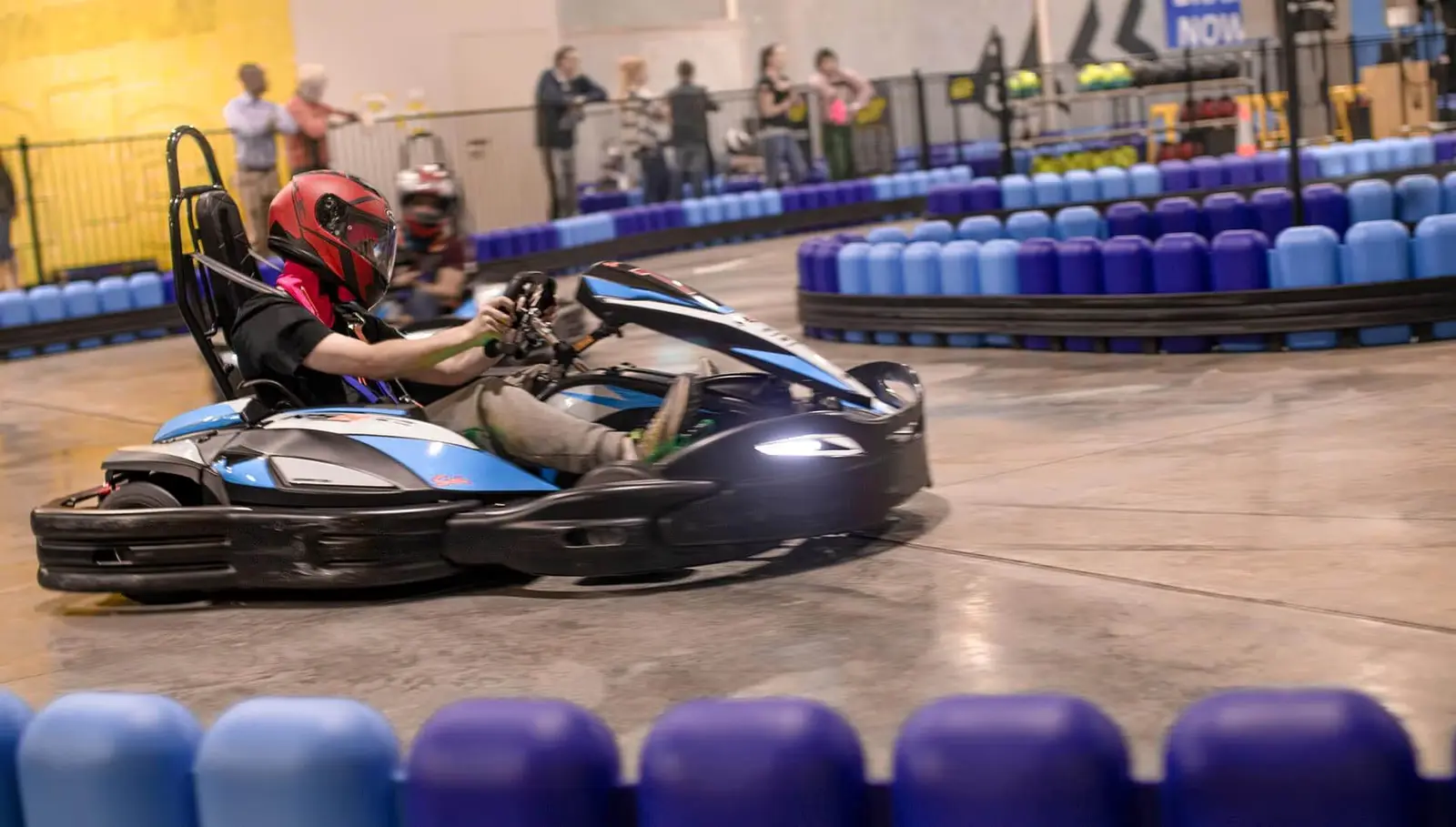 A helmeted PowerPlay racer steering an electric kart around the indoor track